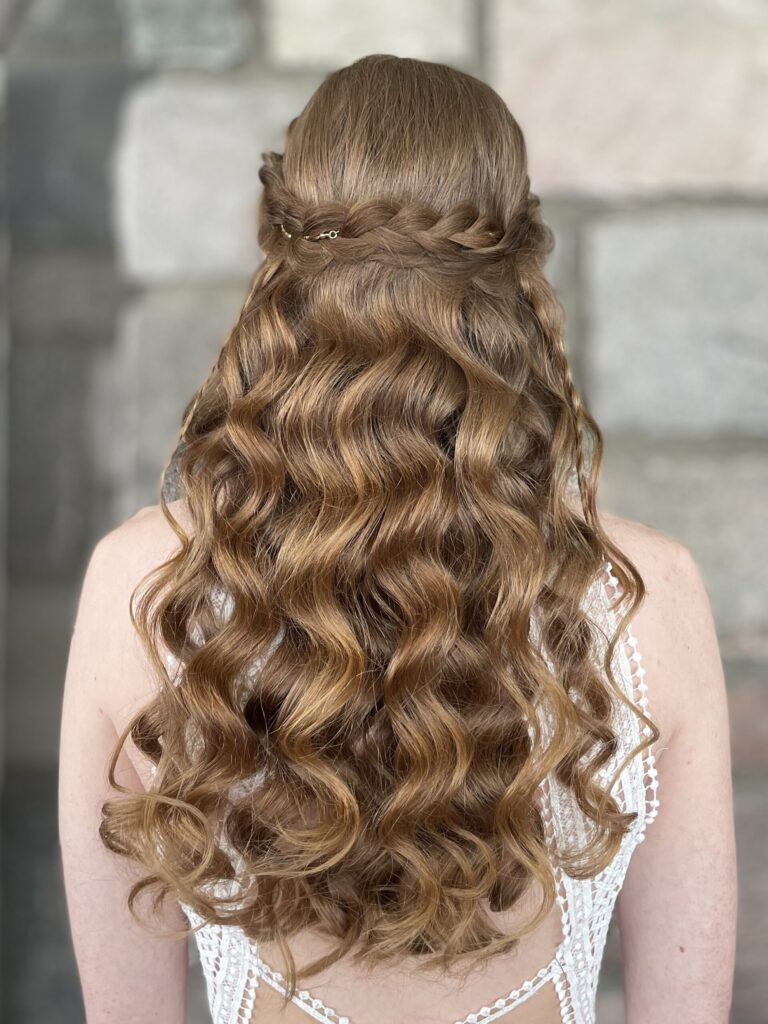 Bride with a half-up half-down style on ginger hair. The hairstyle features a braided section along the crown of the head with ornamental jewelry woven into the main braid, with smaller symmetric braid work framing the curls. two curled face framing pieces are outlining the bride's face. Styled by Belleza Bridal Styles LLC