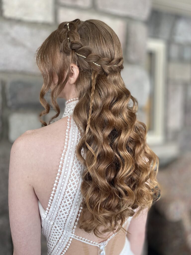 Bride with a half-up half-down style on ginger hair. The hairstyle features a braided section along the crown of the head with ornamental jewelry woven into the main braid, with smaller symmetric braid work framing the curls. two curled face framing pieces are outlining the bride's face. Styled by Belleza Bridal Styles LLC