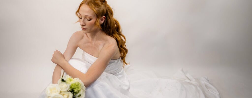Gorgeous woman in white wedding gown, holding a bouquet of green and white flowers, with long flowing curled red-ginger hair in a half-up half-down style with face framing.